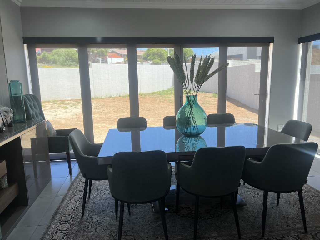 A Simple dining room with grey wide screen roller blinds. A large vase is sitting on the grey dining room table which has upholstered grey dining room chairs to match.