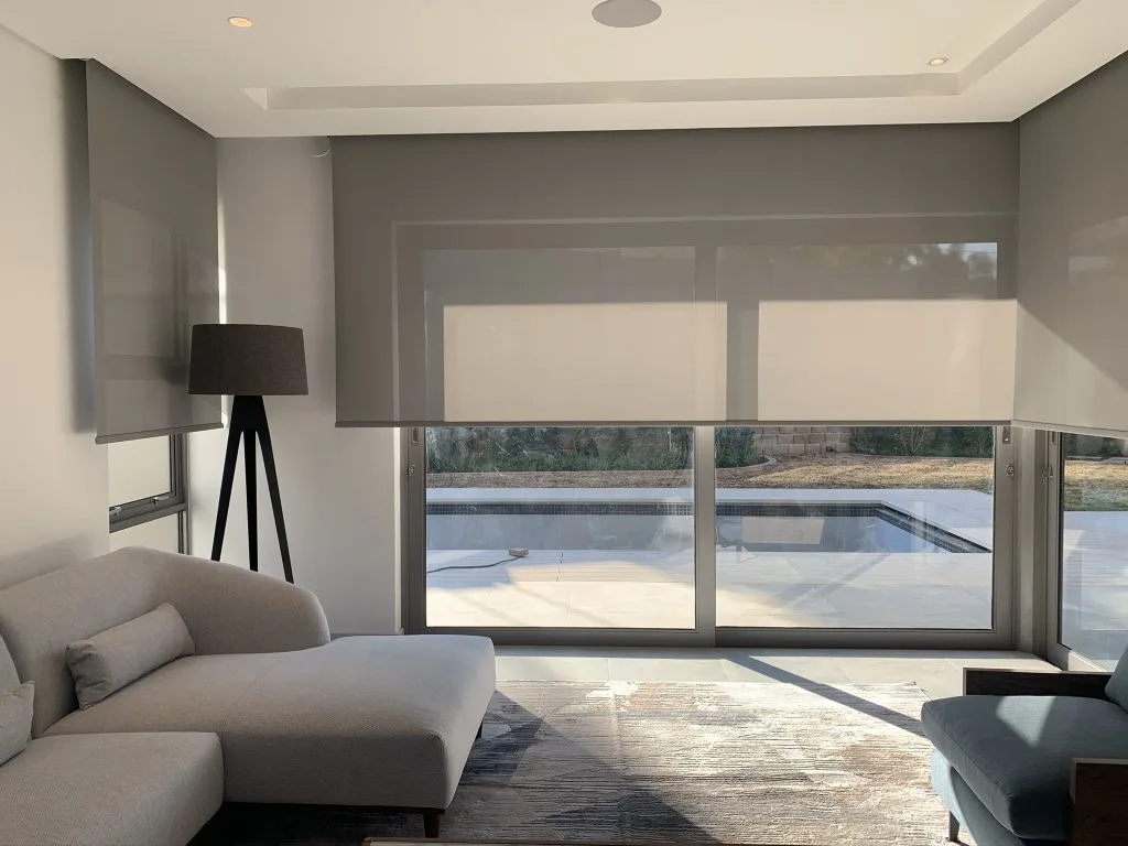 modern living room with grey screen roller blinds showcasing l=its lg=ight filtering effects and creating a warm atmosphere. A light grey couch on the left, modern rug and dark grey lamp in the corner of the room to complete it's modern look. 